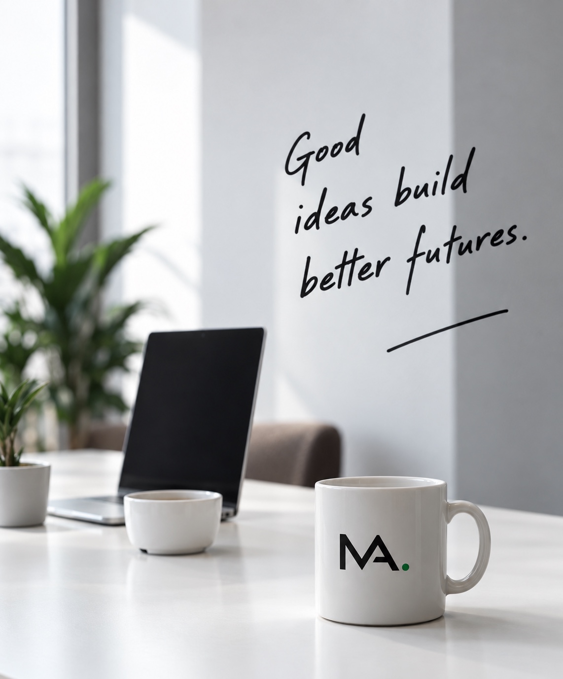 A desk with a laptop, plant and MA mug, beside the words Good ideas build better futures